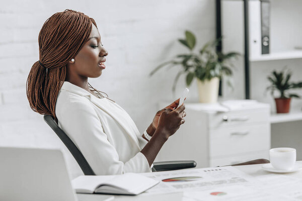side view of confident african american adult businesswoman in white formal wear sitting at desk and using smartphone in office