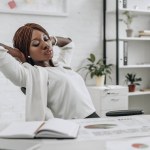 Bela Africano americano adulto empresária em branco formal desgaste com as mãos atrás da cabeça sentado na mesa e relaxante no escritório moderno