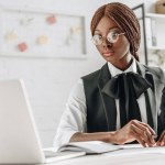 Aantrekkelijke Afro-Amerikaanse volwassen zakenvrouw in glazen zit op computerbureau en werkzaam in kantoor