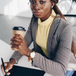 Hermosa mujer de negocios afroamericana seria utilizando gráficos tableta, beber café para ir y mirando la cámara en el lugar de trabajo