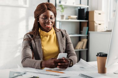 Afrikalı-Amerikalı kadın mimar smartphone kullanıyorsanız ve office planları ile masada çalışıyorsanız gözlük gülümseyen