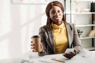 Afrikalı-Amerikalı kadın mimar kahve tutan bardaklarda smartphone kullanarak gidip, Resepsiyon planları ile proje üzerinde çalışan