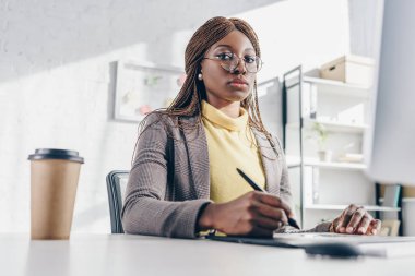 Afrika kökenli Amerikalı yetişkin işkadını bilgisayar masa başında oturan, grafik tablet kullanarak ve işyeri kameraya bakarak konsantre