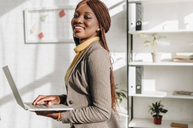 african american adult businesswoman holding laptop at workplace, smiling and looking at camera