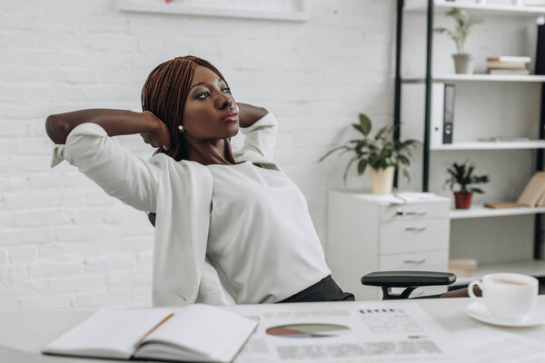 african american adult businesswoman in white formal wear with hands behind head sitting at desk and relaxing at modern office