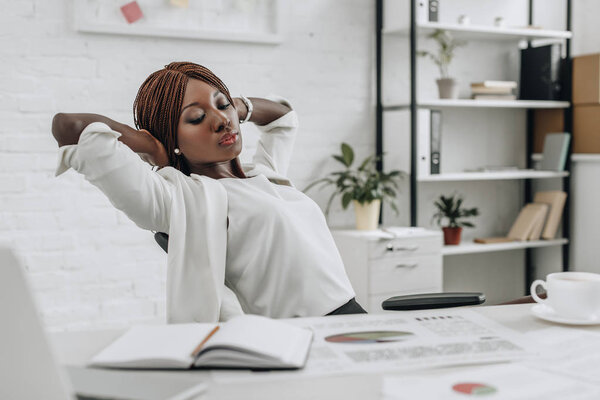 beautiful african american adult businesswoman in white formal wear with hands behind head sitting at desk and relaxing at modern office