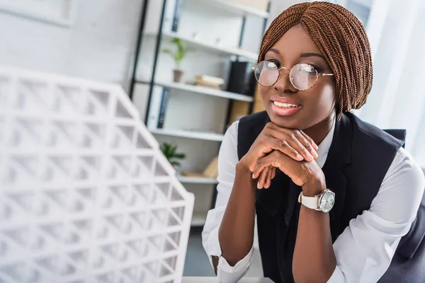 微笑的非洲裔美国成年女建筑师在眼镜和正式穿支持下巴与手 而在办公室的建设项目 — 图库照片