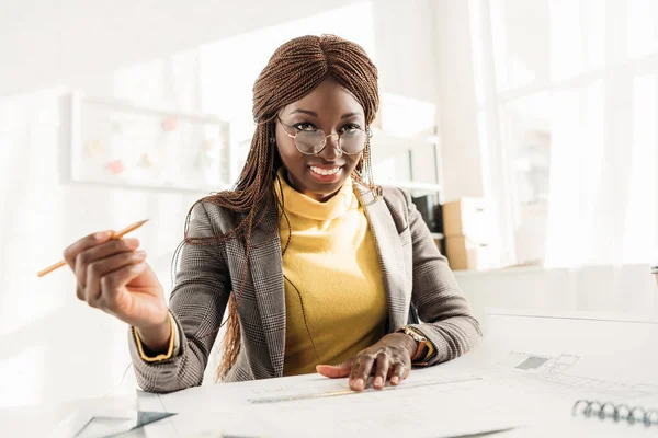 smiling african american female architect in glasses holding pencil, looking at camera and working on project at desk with blueprints in office
