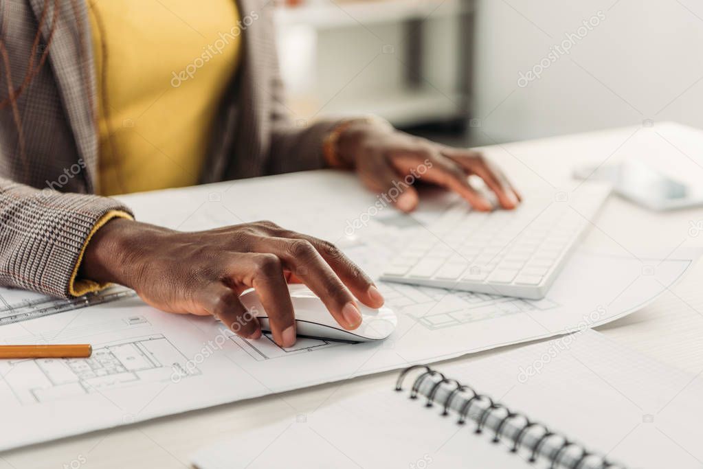 cropped view of african american female architect using compute desk with blueprints
