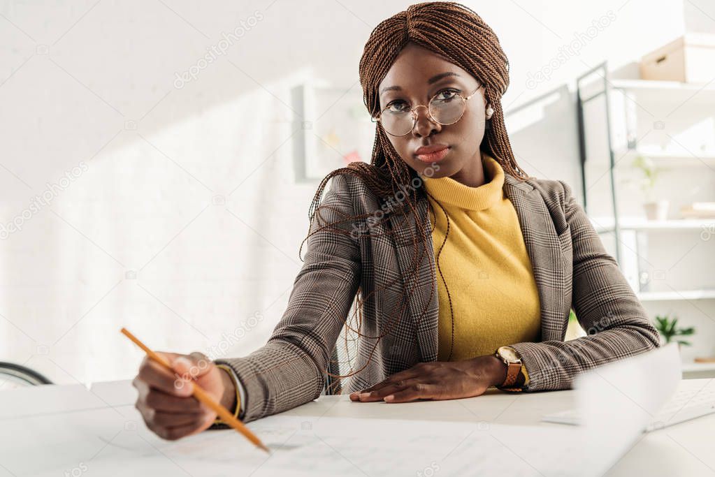 serious african american female architect in glasses holding pen and working on project at desk with blueprints in office