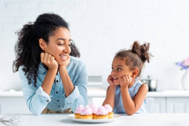 neşeli Afrikalı-Amerikalı anne ve çeneleri ellerde dinlenme ve her diğer, cupcakes mutfakta masada arıyor kızı