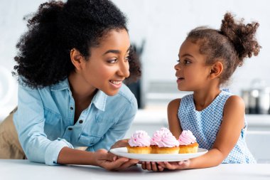 Afrika kökenli Amerikalı anne ve ev yapımı kekler tutarak ve birbirine mutfakta bakarak kız gülümseyerek