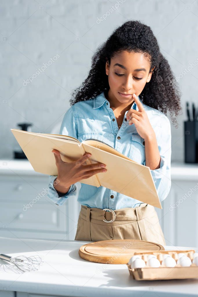 pensive beautiful african american girl reading cookbook in kitchen