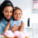 Atractiva africana americana madre abrazando hija en mesa con cupcakes en cocina