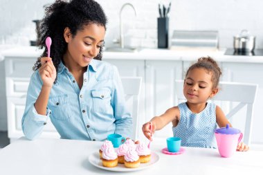 african american mother and daughter eating homemade cupcakes in kitchen clipart