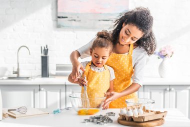 smiling african american mother helping daughter mixing eggs for dough in kitchen clipart