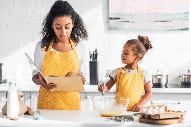 african american mother reading cookbook and daughter preparing dough in kitchen clipart