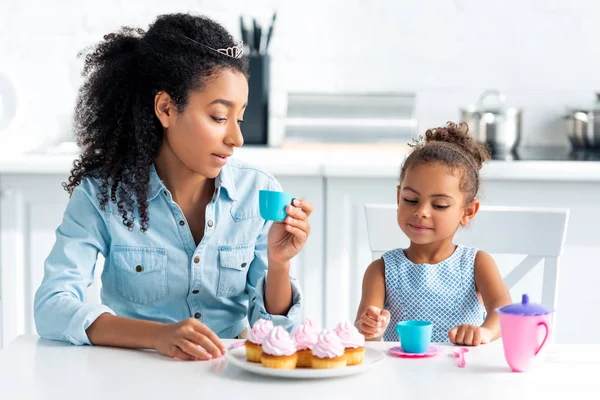 アフリカ系アメリカ人の母と娘は台所でカップケーキとのテーブルに座ってのティアラ  — 無料ストックフォト