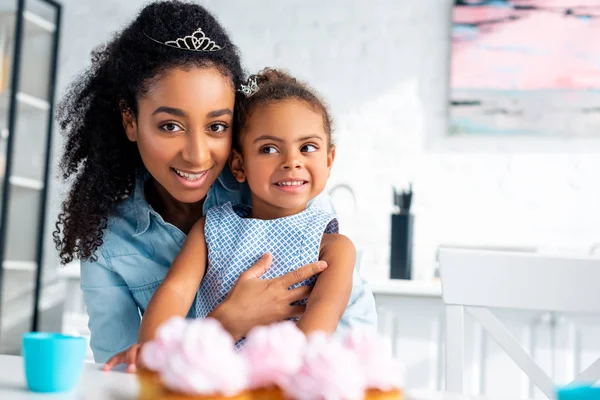 キッチンでカップケーキとテーブルで魅力的なアフリカ系アメリカ人母ハグ娘  — 無料ストックフォト