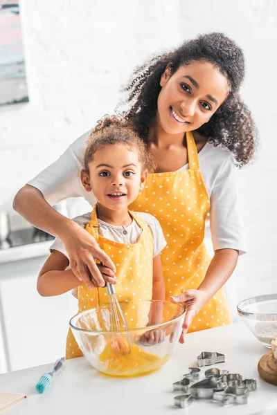 smiling african american mother helping daughter whisking eggs for dough in kitchen, looking at camera