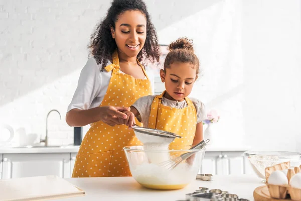 african american mother helping daughter preparing dough and sieving flour in kitchen
