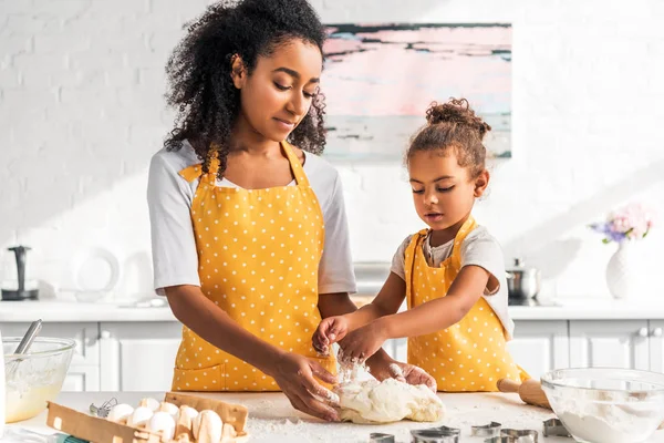 african american mother and daughter in aprons kneading dough with hands in kitchen