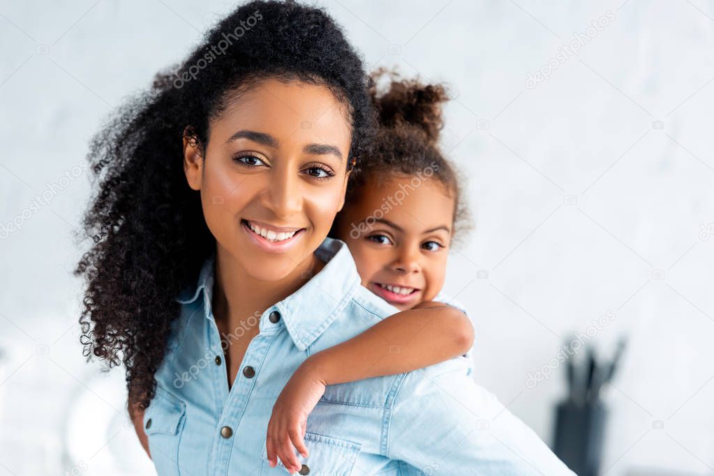 cheerful african american mother giving piggyback to adorable daughter in kitchen