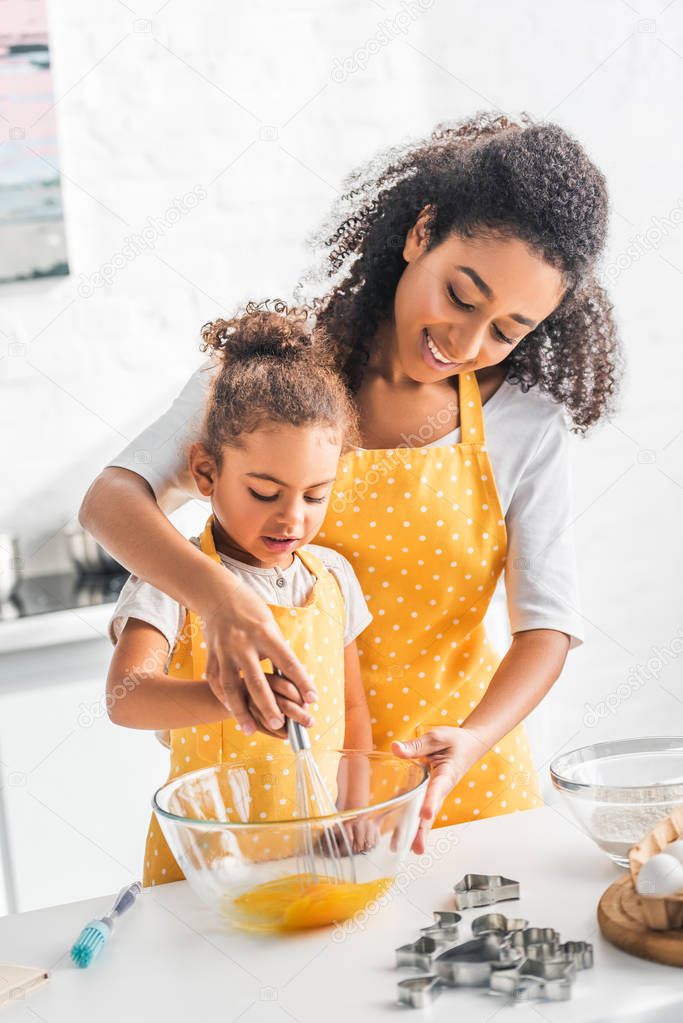 african american mother and daughter whisking eggs for dough together in kitchen