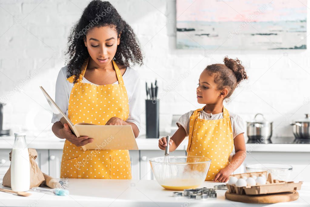 african american mother reading cookbook and daughter preparing dough in kitchen