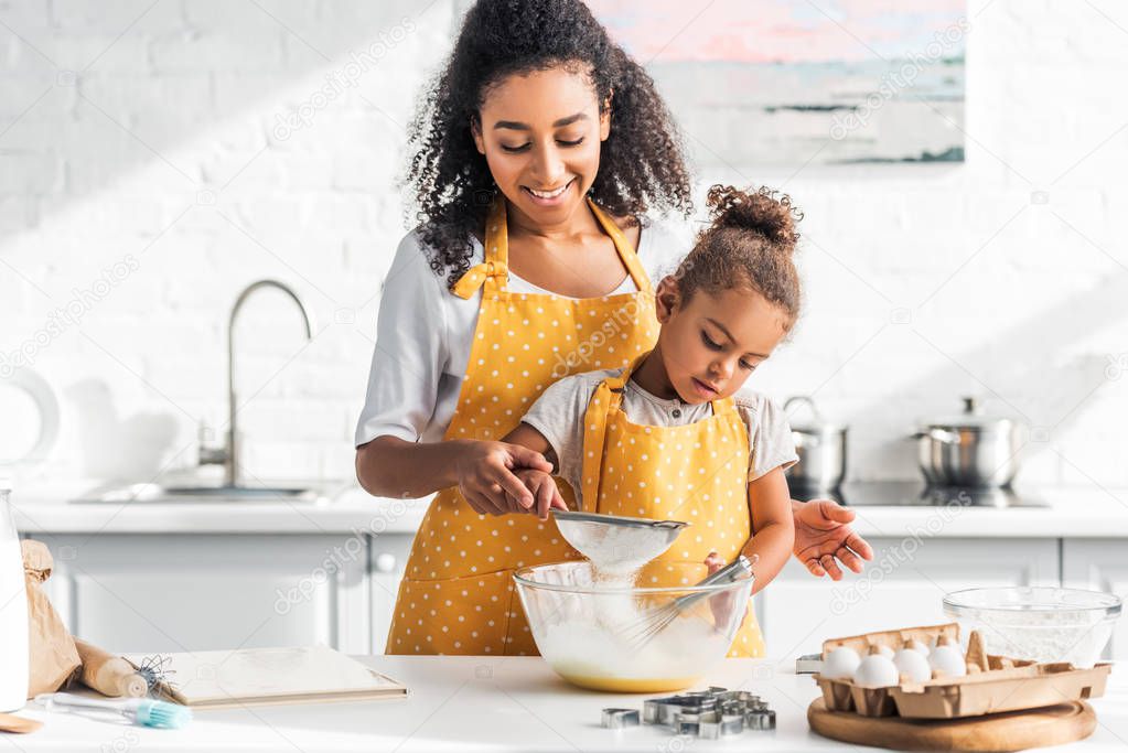 smiling african american mother and daughter preparing dough and sieving flour in kitchen