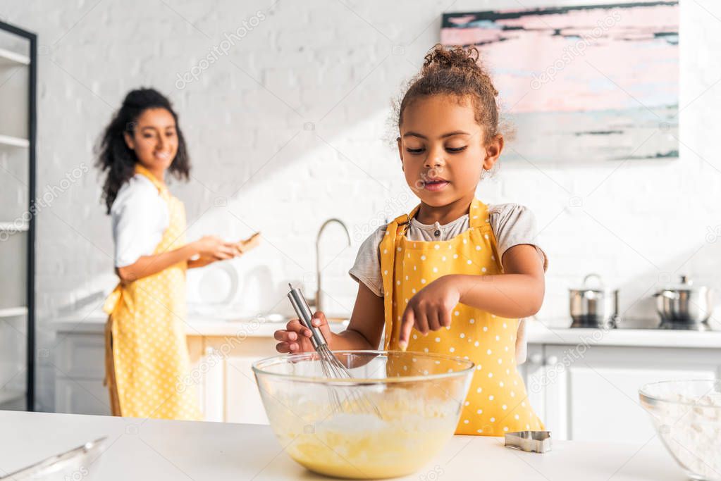 african american daughter preparing and whisking dough, putting finger into bowl in kitchen