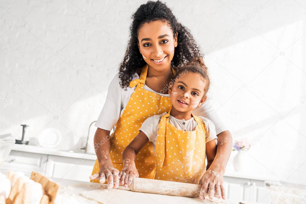cheerful african american mother helping daughter rolling dough with rolling pin in kitchen, looking at camera