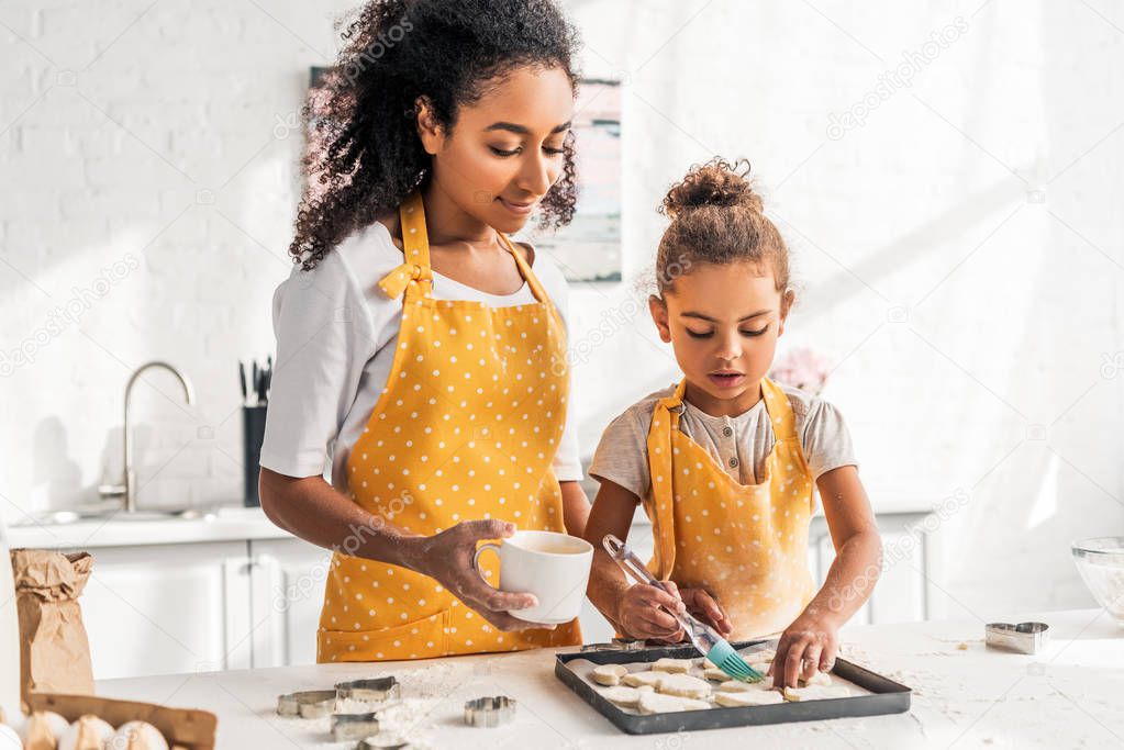 african american mother and daughter in yellow aprons applying oil on unbaked cookies in kitchen