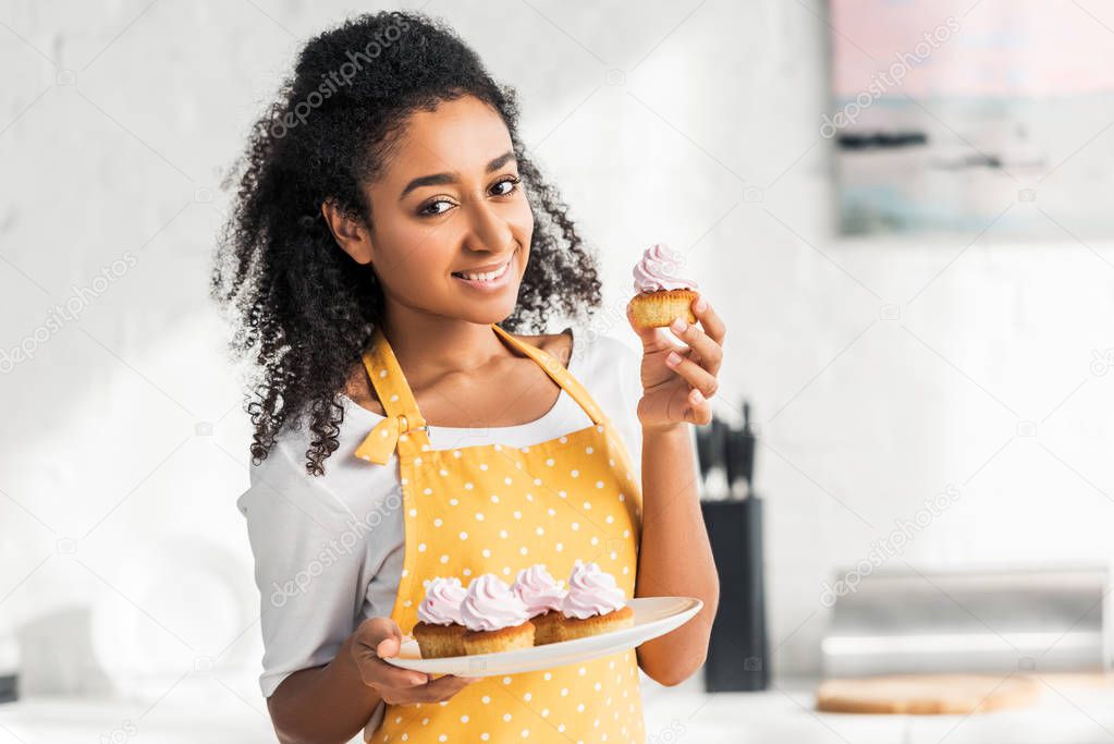smiling attractive african american girl in apron holding homemade cupcakes and looking at camera in kitchen