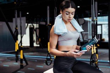 Spor salonunda spor şişe tutarak akıllı telefon kullanan çekici sportif kız 