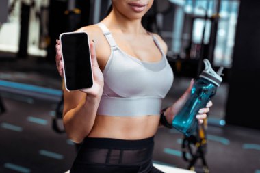 Fitness salonunda smartphone ve spor şişe tutan sportif kadın kırpılmış görünümünü 