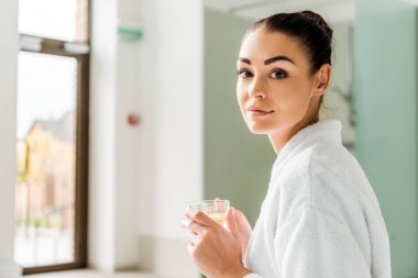 Bitkisel içecek fincan tutarak ve spa kamerada bakarak bornoz çekici genç kadın