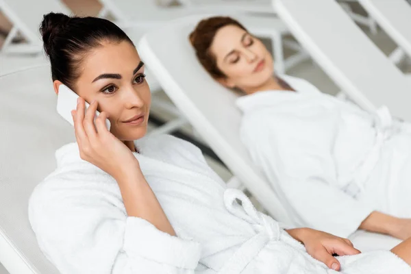 微笑的年轻女子在浴袍交谈的智能手机 而休息在水疗中心的日光浴床 — 图库照片
