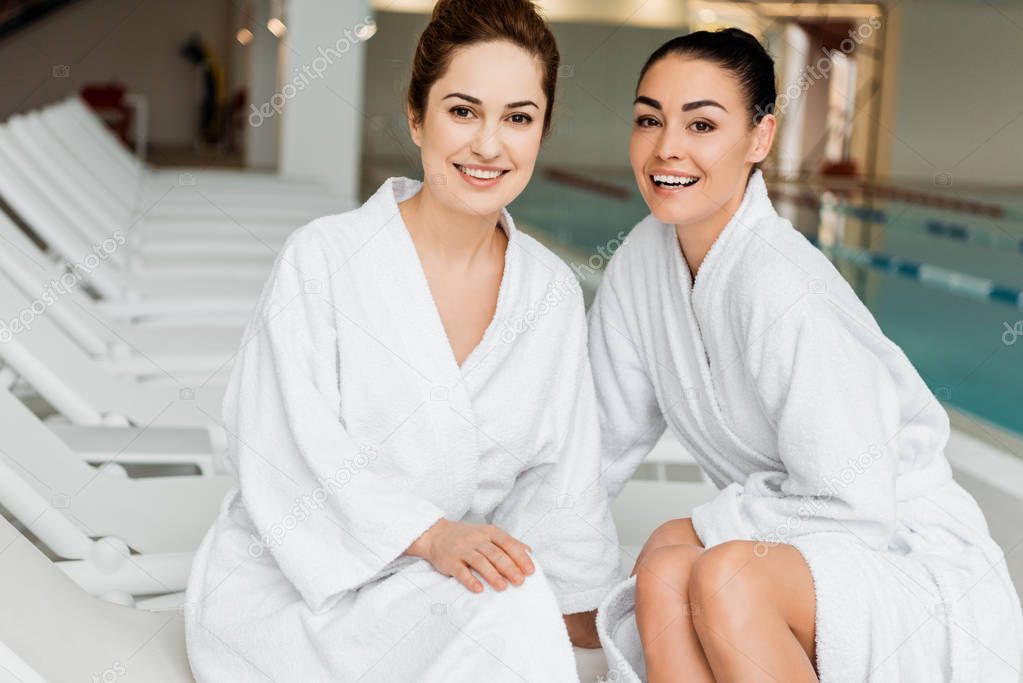 happy young women in bathrobes smiling at camera while relaxing together in spa