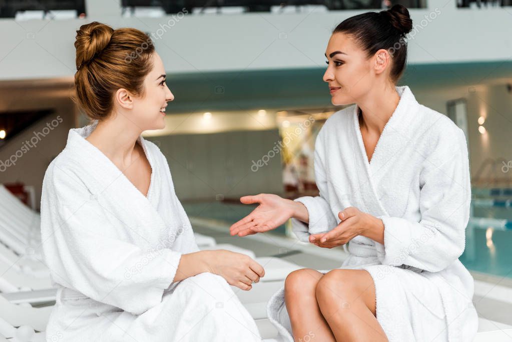 smiling young women in bathrobes talking while relaxing together in spa