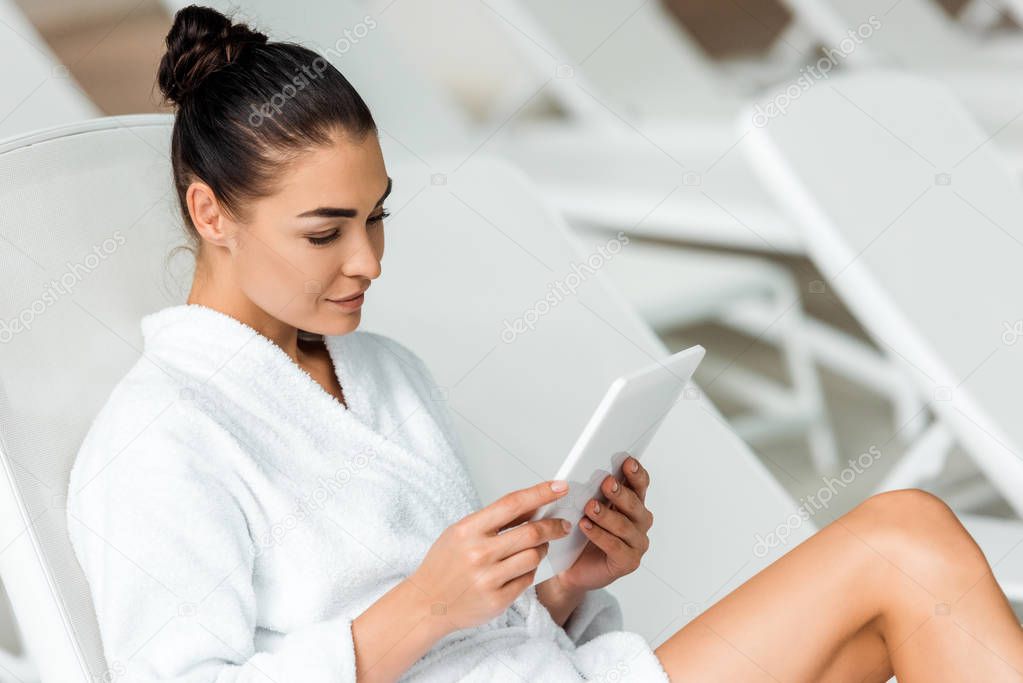 attractive young woman in bathrobe using digital tablet on sunbed in spa 