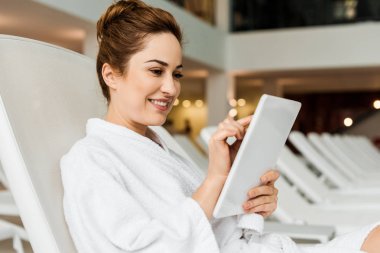 Spada dinlenirken dijital tablet kullanarak bornoz gülümseyen genç kadın  