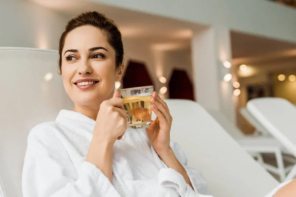 beautiful smiling young woman in bathrobe holding cup with herbal drink while relaxing in spa