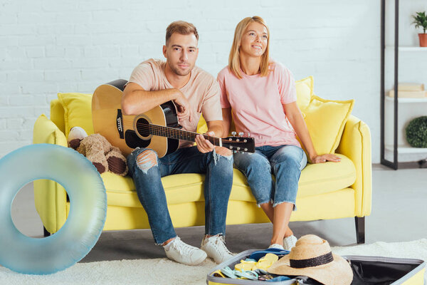husband holding guitar while wife dreaming about summer holidays and sitting on couch at home