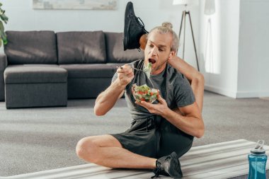 yoga mat üzerinde oturan ve sebze salata yemek başının arkasında bacak ile esnek yetişkin sporcu  