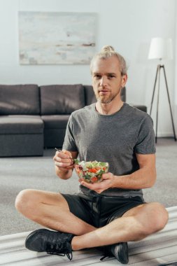 atletik erkekte tabak sebze salatası tutarak ve yoga mat evde otururken kameraya seyir spor giyim 