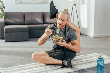 yoga mat üzerinde oturan ve sebze salata yemek başının arkasında bacak ile esnek yetişkin adam 