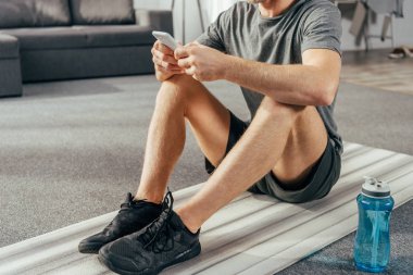 cropped shot of athletic man in sportswear sitting on yoga mat and using smartphone at home clipart