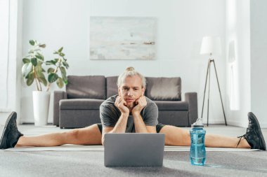 sporty adult man doing split on yoga mat and looking at laptop