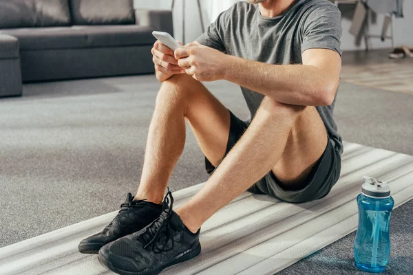 运动男子在运动服装坐在瑜伽垫上 并在家里使用智能手机的裁剪镜头 — 图库照片
